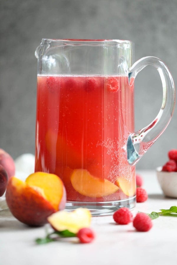 A tall glass pitcher of Raspberry-Peach Rosé Sangria with a grey backdrop. 