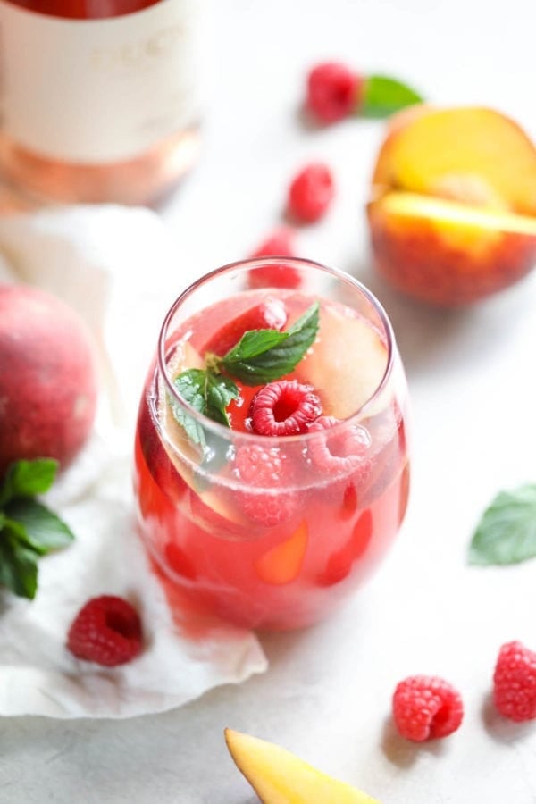 A glass of summer awaits with this Raspberry-Peach Rosé Sangria, shown here on a white table surrounded by fresh fruit.