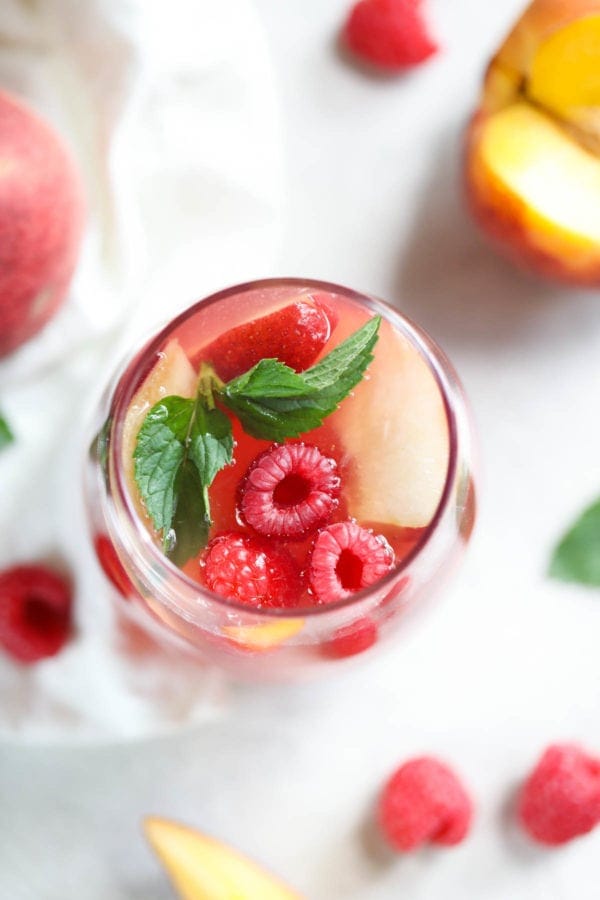 An overhead view of a glass of Raspberry-Peach Rosé Sangria with peach slices, three plump berries, and a sprig of mint floating at the top.