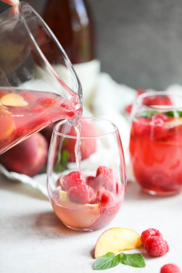 A glass of Raspberry-Peach Rosé Sangria is poured from a pitcher into a stemless wine glass filled with fresh raspberries and peach slices.