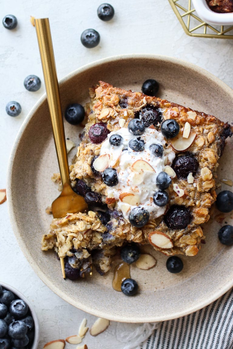 Overhead view piece of blueberry baked oatmeal on stone plate topped with yogurt, almonds, and fresh blueberries.