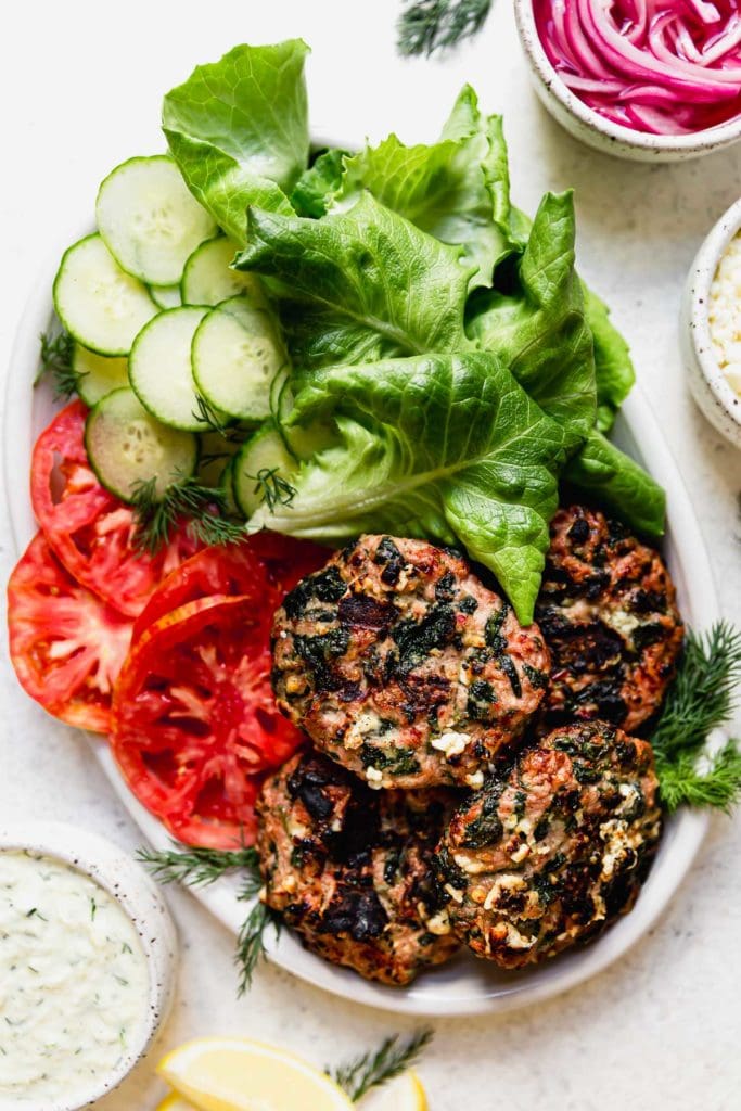 Large white platter holding the Grilled Turkey Burgers, tomato slices, lettuce leaf, cucumber slices and dill. 