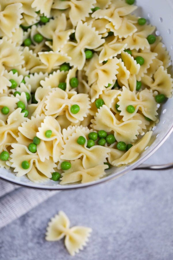 Bow tie pasta in a white colander with green peas scattered throughout to make tuna pasta salad.