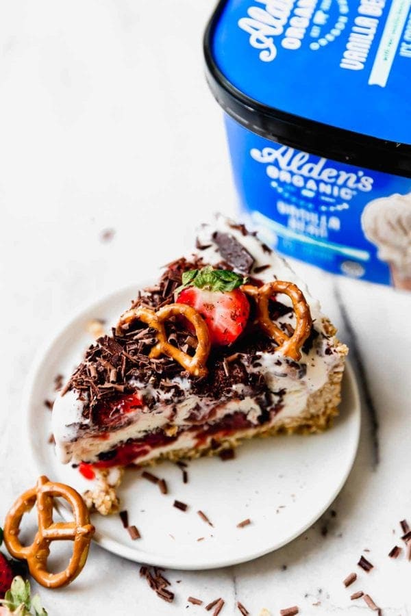 A slice of Strawberry Pretzel Ice Cream Pie with shaved chocolate, pretzels, and fresh strawberry sits on a small white plate in front of a blue tub of Alden's Organic Vanilla Bean Ice Cream.