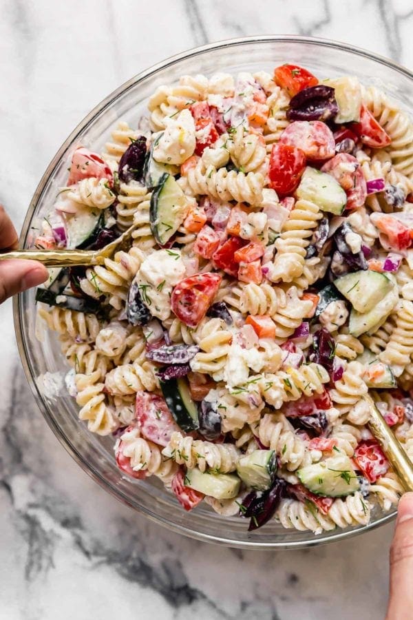 Greek gluten-free veggie pasta salad in a glass bowl being mixed with two gold spoons.