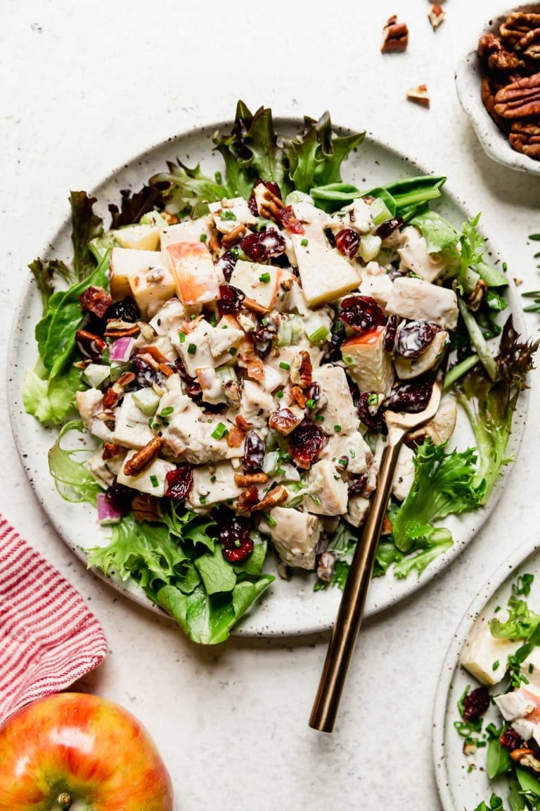 Overhead view of a bed of greens topped with cranberry chicken salad with apples and topped with dried cranberries, toasted pecans, crisp bacon, fresh rosemary, and creamy mayo. 