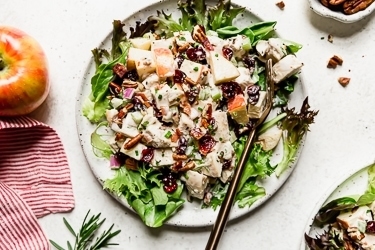 Creamy chicken salad topped with toasted pecans and dried cranberries on a bed of mixed greens on cream speckled plate