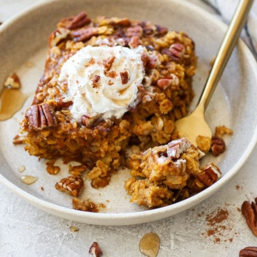 Gluten-free pumpkin baked oatmeal on a cream plate with a gold fork and topped with whipped cream and pecans.