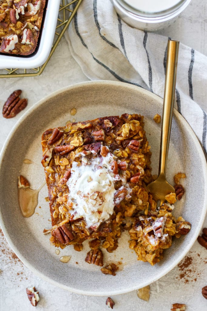 Serving of Pumpkin Baked Oatmeal on a light-colored plate topped with whipped topping, maple syrup and chopped pecans. Gold fork also on the plate holding a bite.