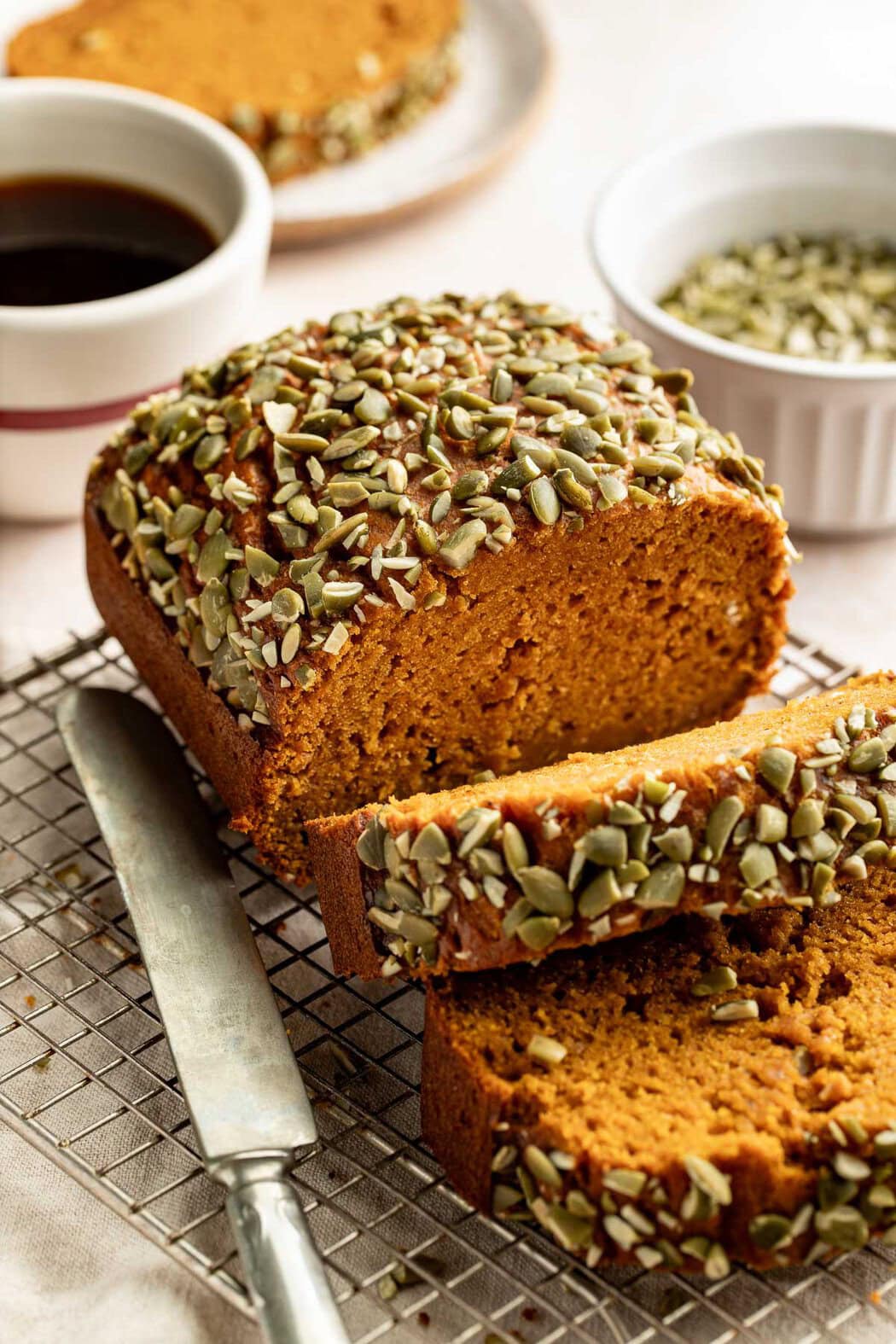 Slices of a loaf of pumpkin bread topped with crushed pepitas topple forward on a wire rack.