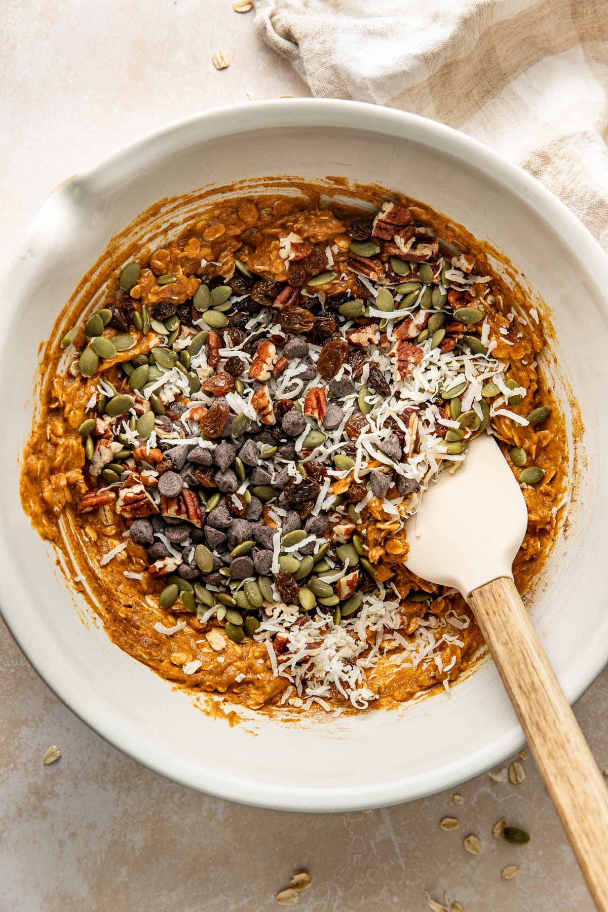 Chocolate chips, coconut flakes, chopped pecans, and pepitas being folded into pumpkin breakfast cookie batter.