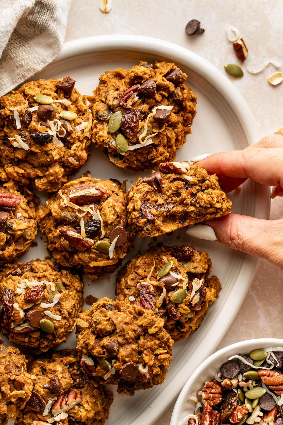 A hand holding a pumpkin breakfast cookie missing a bite next to a platter of breakfast cookies.