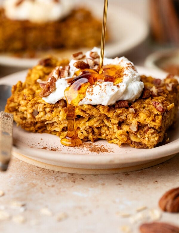 Side view of slice of pumpkin baked oatmeal on plate topped with yogurt, maple syrup being drizzled over top