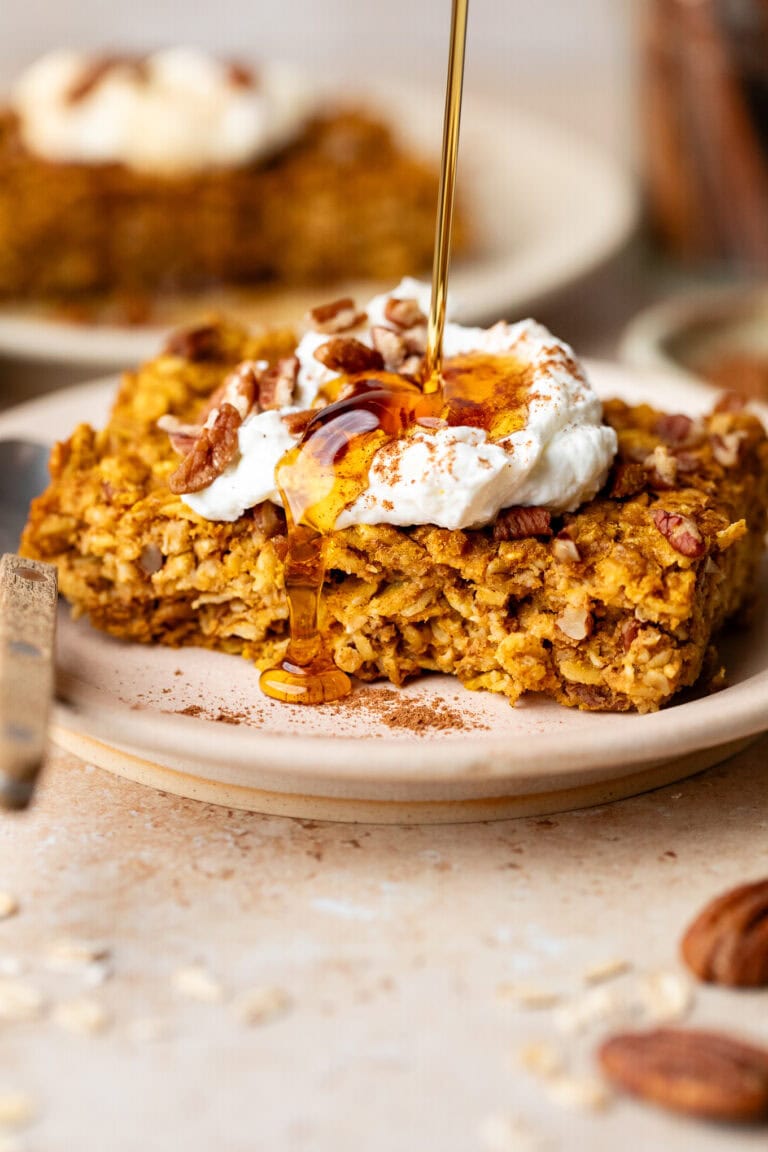 A slice of pumpkin baked oatmeal on plate topped with yogurt, maple syrup being drizzled over top.
