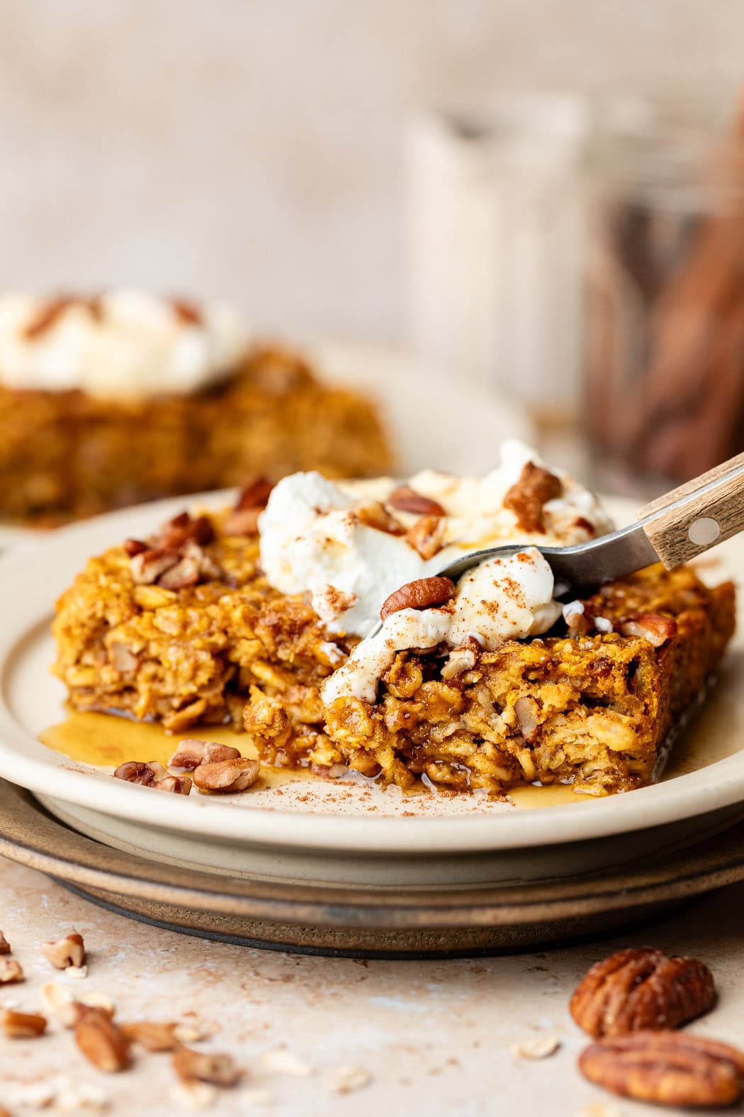 A spoon scooping a bite of a freshly baked piece of Pumpkin Baked Oatmeal topped with whipped cream and drizzled with maple syrup. 