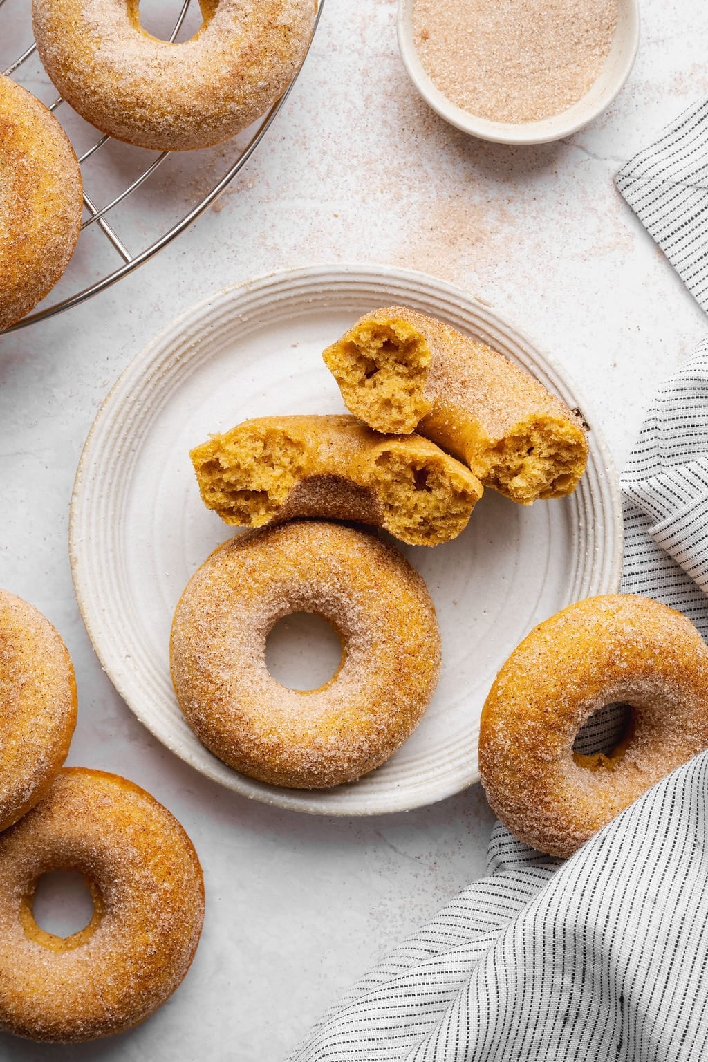 A plate of two pumpkin donuts, one whole and one broken in half to show the inside texture with other donuts nearby on a wire cooling rack.