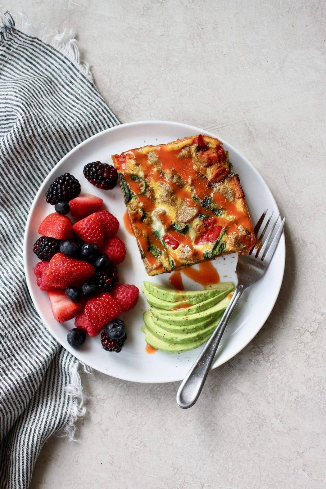 Egg bake with hot sauce drizzled on top, fresh berries, and slices of avocado on a white plate