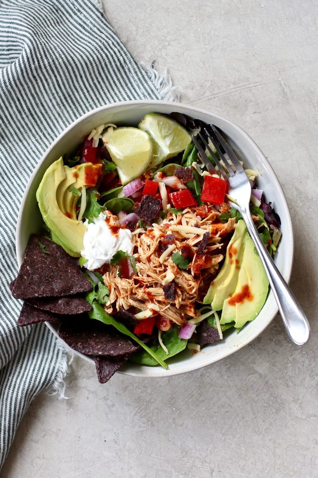 Shredded chicken over a mixed green salad and topped with avocado, lime, and blue chips in a white bowl