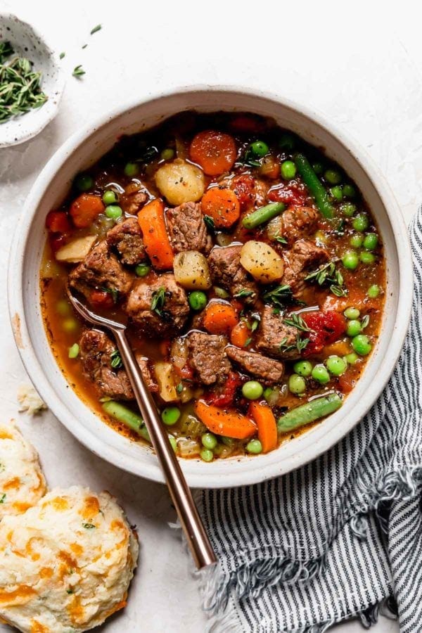 Overhead view of vegetable beef soup with stew meat, potatoes, and veggies served in a cream bowl with biscuits on the side.