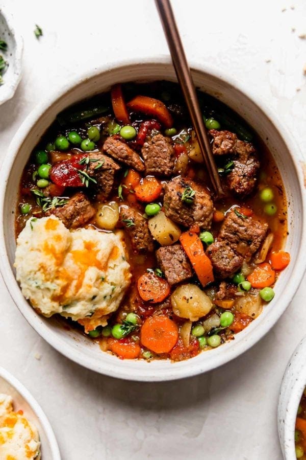 A big bowl filled with hearty vegetable beef soup and topped with cracked pepper and fresh thyme with a cheddar biscuit on the side