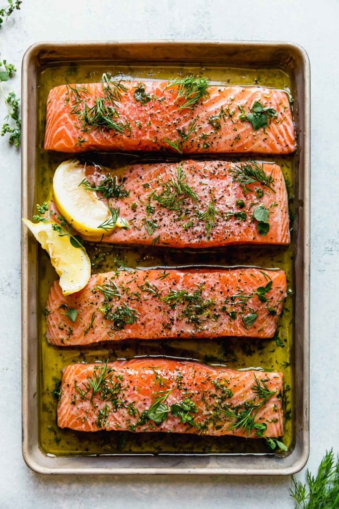Four salmon fillets for the Mediterranean bowl. The salmon fillets are on a sheet pan marinating in a lemon-herb marinade and topped with fresh herbs and a couple lemon wedges. 