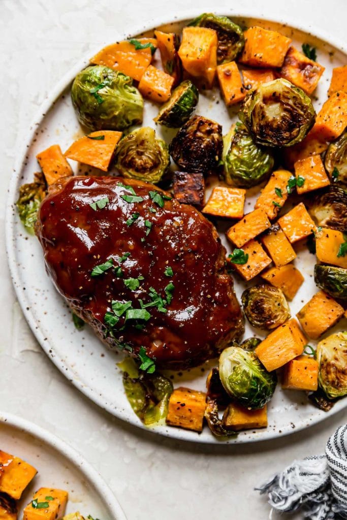 Mini meatloaf topped with barbecue sauce on a plate with roasted sweet potatoes and Brussels sprouts. 