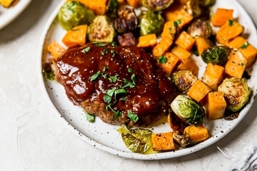 A mini meatloaf with roasted sweet potatoes and brussels sprouts on a speckled plate