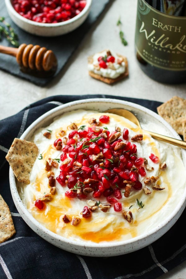 Honey Whipped Goat Cheese with Pomegranate in white serving bowl with a gold spoon topped with fresh thyme and honey. Served with crackers.