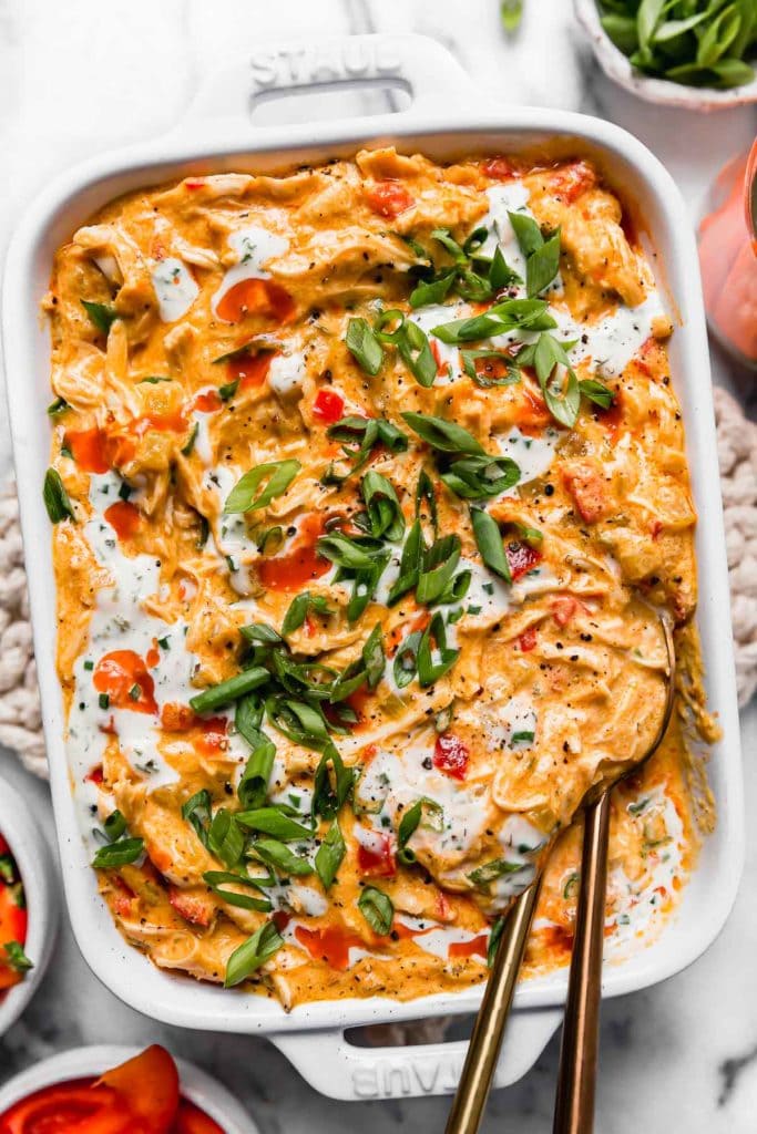 Crockpot Buffalo Chicken Dip in a white casserole dish garnished with green onion and a drizzle of ranch. Two gold spoons in the dish for serving.