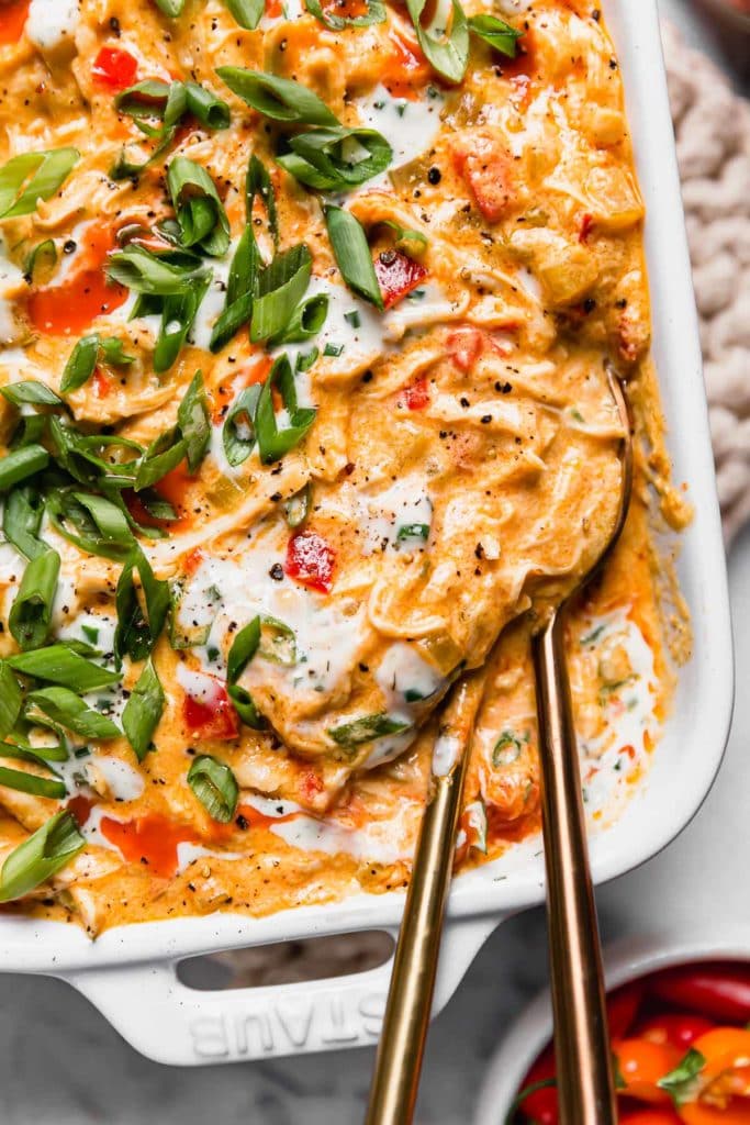 Crockpot Buffalo Chicken Dip in a white casserole dish garnished with green onion and a drizzle of ranch. Two gold spoons in the dish for serving.