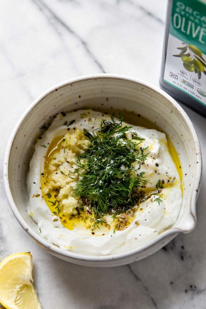 A small white, speckled bowl filled with ingredients for the yogurt sauce including yogurt, lemon juice, lemon zest, garlic, fresh dill and black pepper.