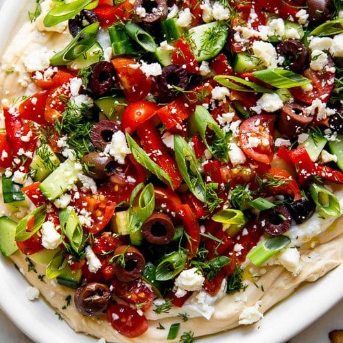 A layered Greek-inspired hummus dip topped with cucumber, kalamata olives, cherry tomatoes, and fresh dill.