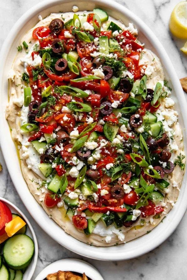 Greek Hummus Dip served on a large, white platter. A thick layer of hummus topped with a layer of Tzatziki, cucumber, tomatoes, feta, olives, green onion, herbs and drizzled with olive oil.