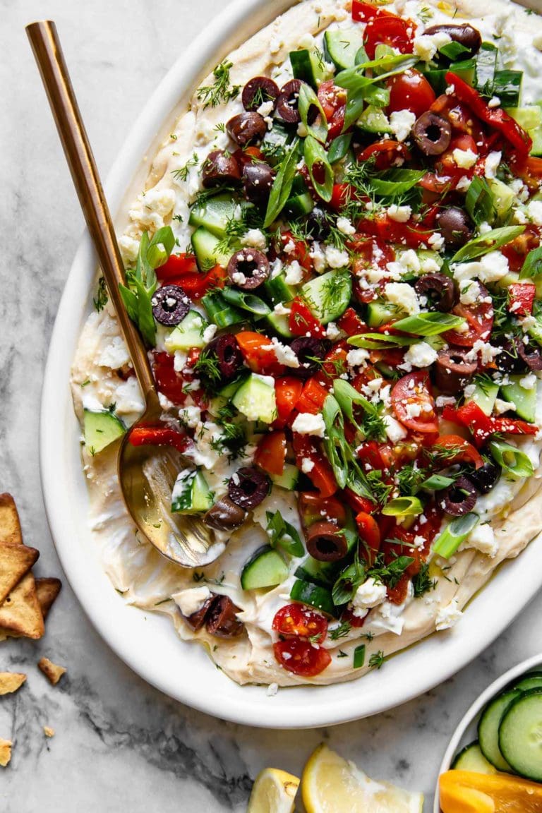 Overhead view layered Greek hummus on a white platter topped with chopped cucumber, kalamata olives, tomatoes, and green onions