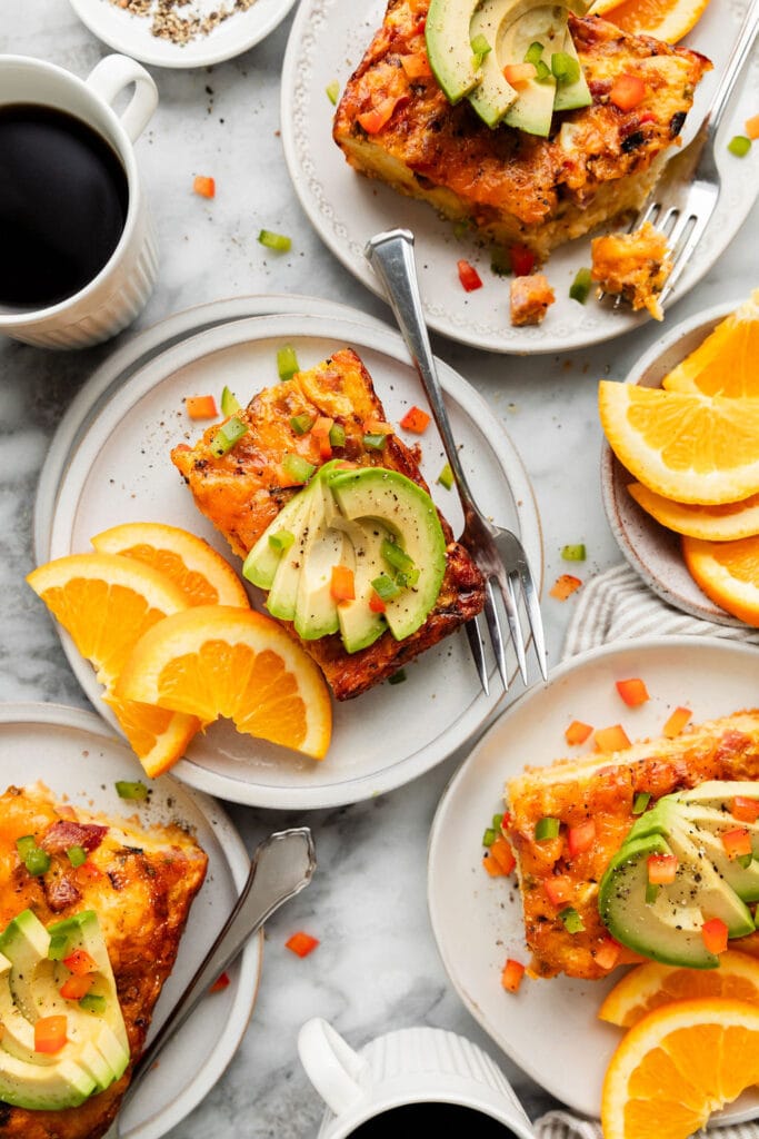 Several plates with servings of breakfast casserole topped with sliced avocado and orange slices on the side; cups of coffee around the plates