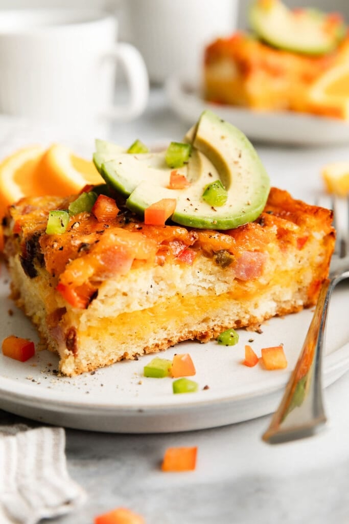 A single serving of ham and cheese breakfast casserole on a plate topped with avocado slices and diced green and red peppers