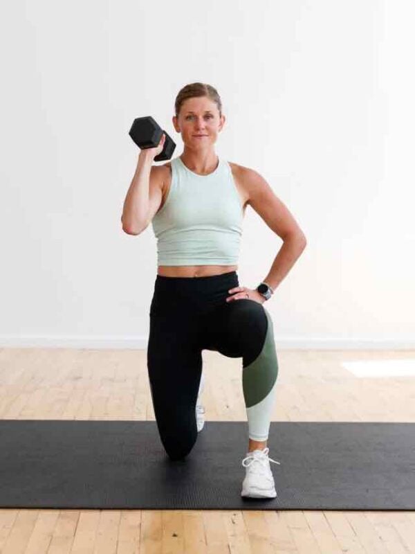 A woman in workout clothes kneeling on one knee holding dumbbell by shoulder for a workout
