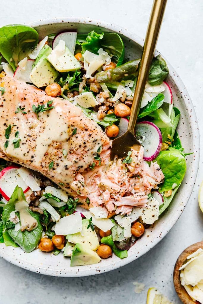 Caesar Salmon Salad Grain Bowl in a speckled stone bowl with greens, avocado, crispy chickpeas and flaky salmon.