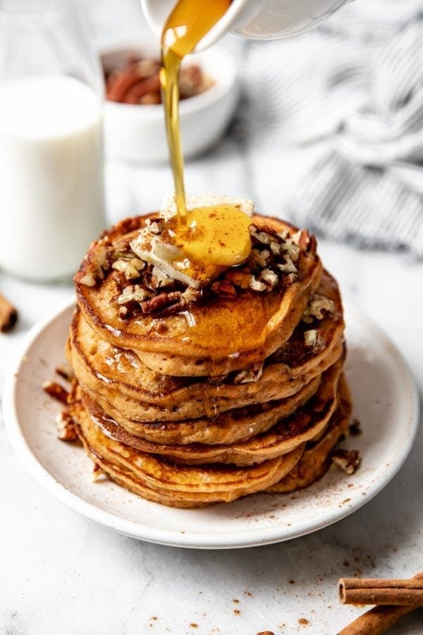 A tall stack of sweet potato pancakes on a white plate topped with a pad of butter, pecans, and maple syrup.