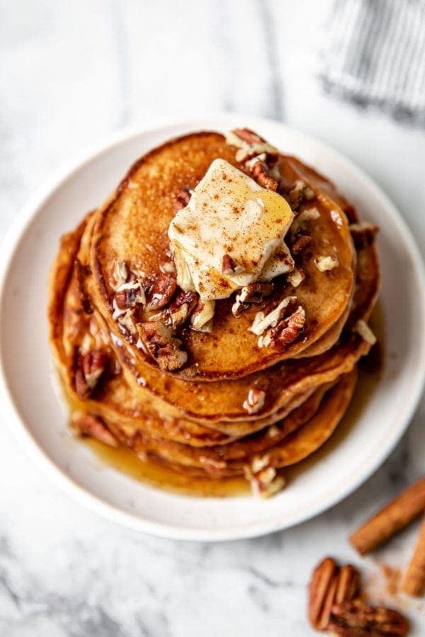 Overhead view of a stack of sweet potato gluten free pancakes on a white plate topped with butter, pecans, and maple syrup.