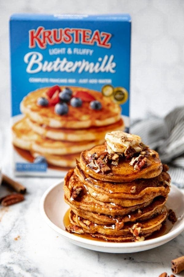 A stack of gluten free sweet potato pancakes on a white plate in the forefront and a blue box of Krusteaz pancake mix in the background