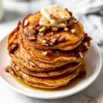 A large stack of sweet potato pancakes on a white plate topped with a pad of butter, pecans, and maple syrup.