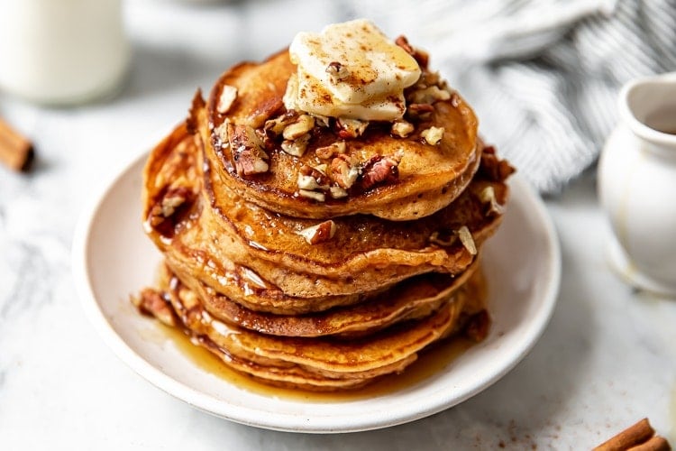Tall stack of fluffy sweet potato pancakes on white plate topped with butter slices and pecans.