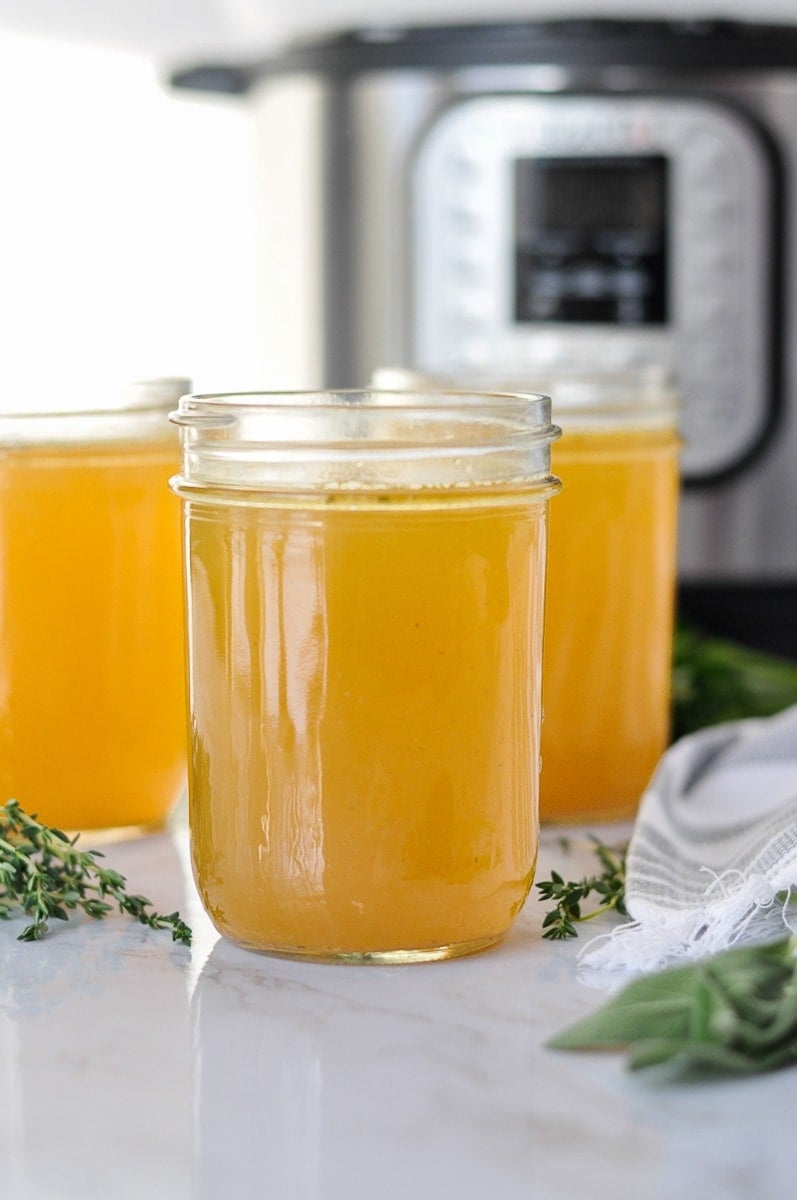 Three mason jars filled with Instant Pot bone broth with fresh herbs scattered around them