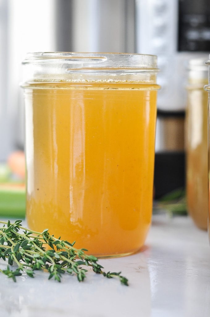 A glass mason jar filled with homemade chicken bone broth with a sprig of rosemary nearby