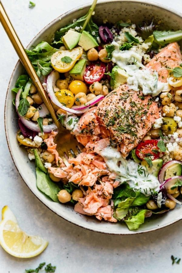 Mediterranean Salmon salad served in a speckled bowl over greens and topped with tzatziki sauce, chickpeas, and dill weed