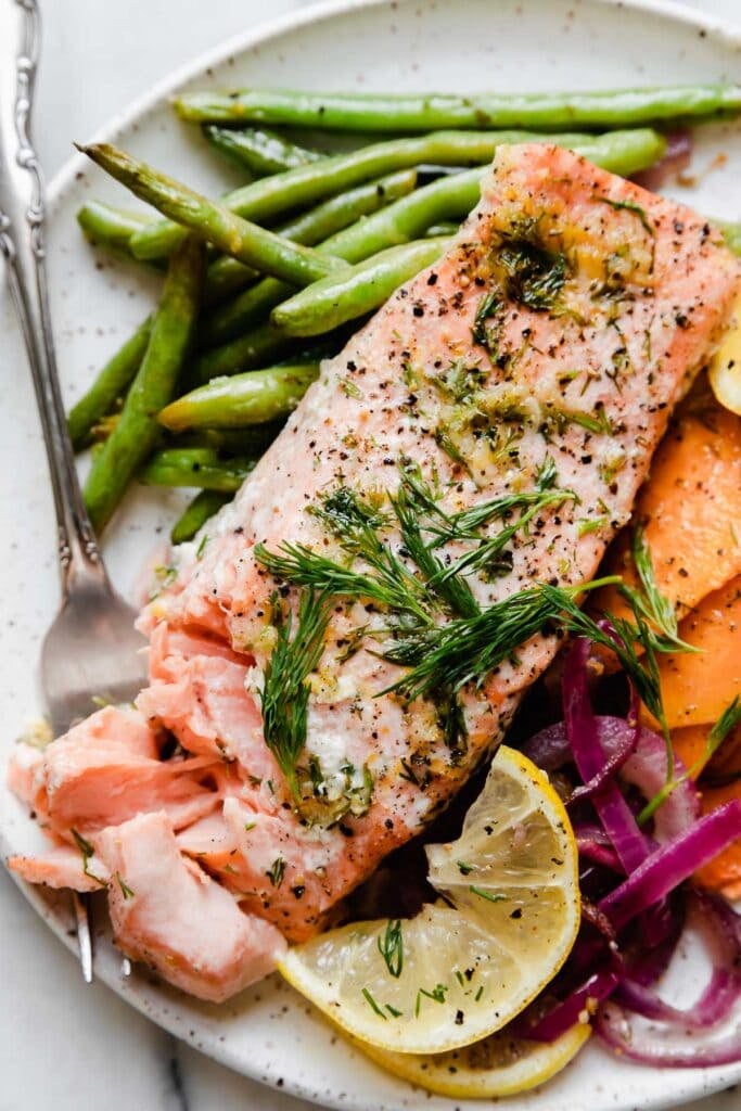 A salmon fillet plated with green beans, purple onion, and sweet potato rounds with a forkful of flakey salmon on the side.