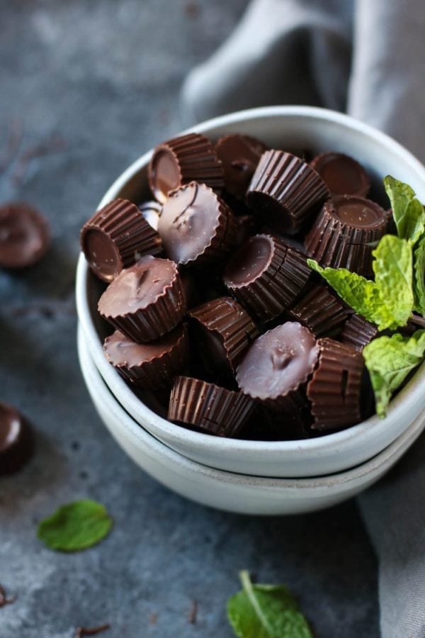 Two white bowls stacked up, top bowl filled to overflowing with dark chocolate mint cups, fresh mint sprigs as garnish. 