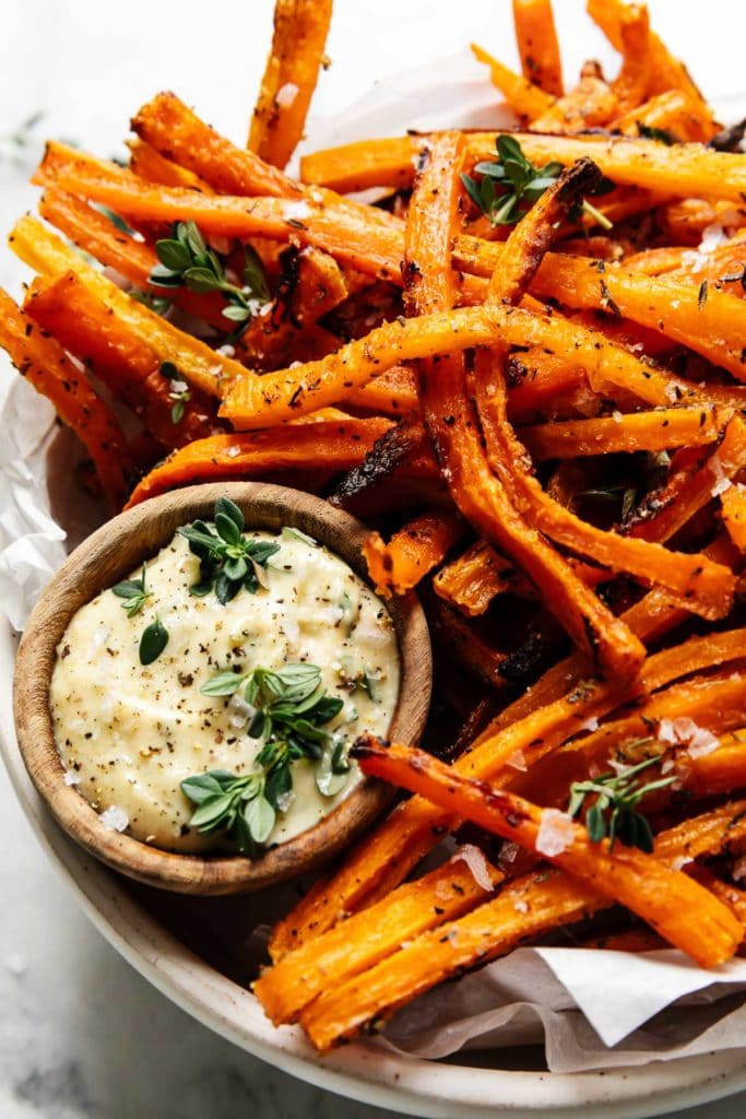 Baked carrot fries in a speckled bowl sprinkled with thyme and sea salt flakes with a small wooden bowl filled with dijon aioli