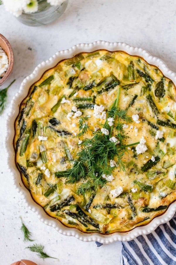 Crustless asparagus, leek, and feta quiche in a white pie plate topped with crumbled feta cheese and fresh herbs.